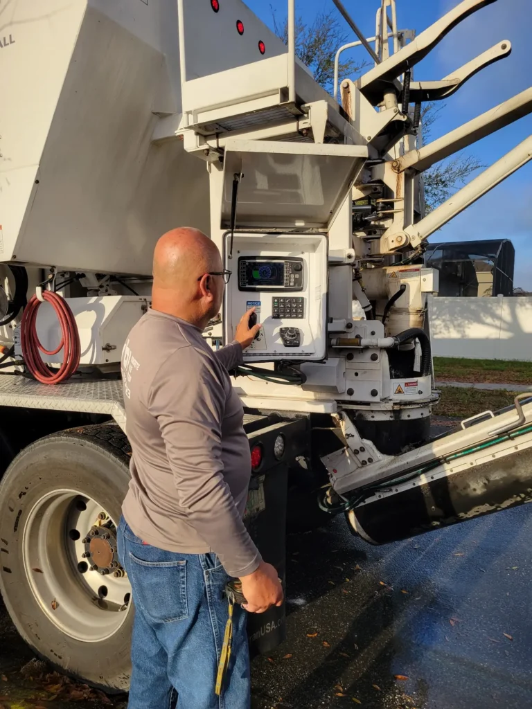 A man operates a control panel on the side of a large pay-by-the-yard concrete delivery truck outdoors. He is wearing glasses, a gray shirt, and blue jeans, standing on wet pavement with sunlight shining on the scene.